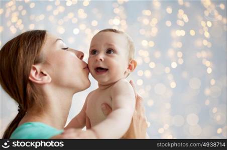 family, child and parenthood concept - close up of happy smiling young mother kissing little baby over lights background. happy young mother kissing little baby over lights