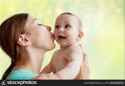 family, child and parenthood concept - close up of happy smiling young mother kissing little baby over summer green lights background. happy young mother kissing little baby