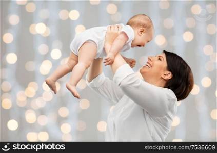 family, child and parenthood concept - happy smiling middle-aged mother holding little baby daughter over festive lights background. happy middle-aged mother with baby over lights