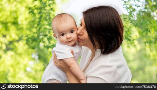 family, child and parenthood concept - happy smiling middle-aged mother kissing little baby daughter over green natural background. happy mother kissing little baby daughter