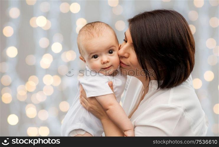family, child and parenthood concept - happy smiling middle-aged mother kissing little baby daughter over festive lights background. happy mother kissing little baby over lights