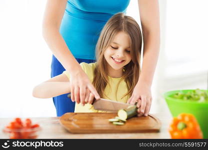 family, child, cooking and home concept - smiling little girl with mother chopping cucumber in the kitchen