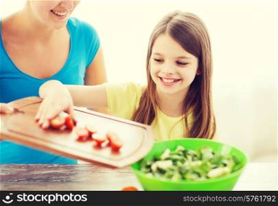 family, child, cooking and home concept - smiling little girl with mother adding tomatoes to salad in the kitchen