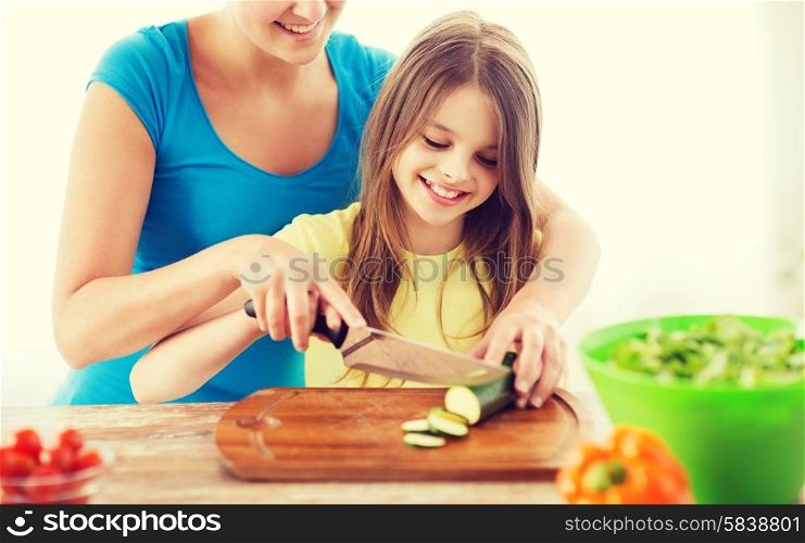 family, child, cooking and home concept - smiling little girl with mother chopping cucumber in the kitchen
