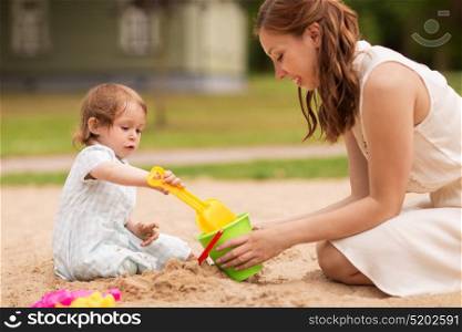 family, childhood and parenthood concept - happy mother and little baby girl with sand toys playing in sandbox at summer playground. happy mother with baby girl playing in sandbox