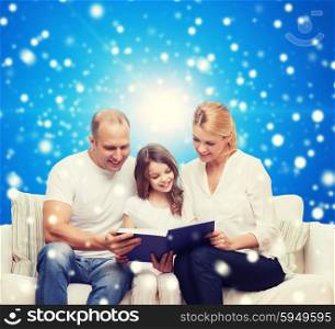 family, childhood, christmas holidays and people - smiling mother, father and little girl reading book over blue snowy background