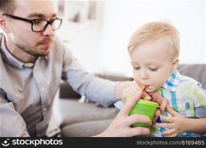 family, childhood, fatherhood, care and people concept - father helping little son with drinking from cup at home