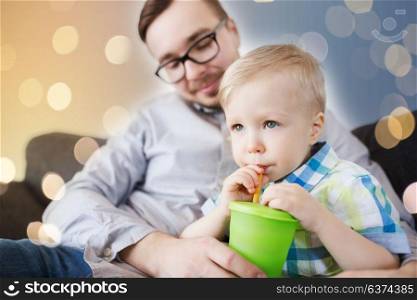 family, childhood, fatherhood, care and people concept - father helping little son with drinking from cup at home. father and son drinking from cup at home