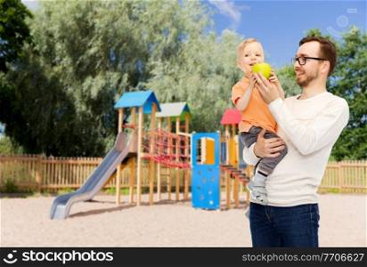 family, childhood, fatherhood, healthy eating and people concept - happy father and and little son with green apple over children’s playground background. happy father and little son with green apple