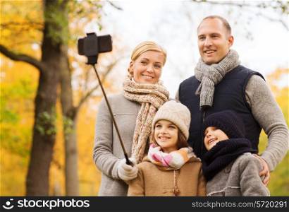 family, childhood, season, technology and people concept - happy family photographing with smartphone and selfie stick in autumn park