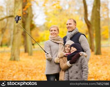 family, childhood, season, technology and people concept - happy family taking selfie with smartphone and monopod in autumn park. happy family with smartphone and monopod in park