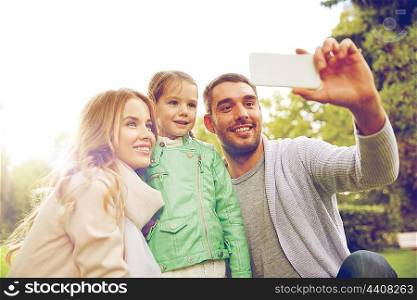 family, childhood, technology and people concept - happy father, mother and little daughter taking selfie by smartphone in park
