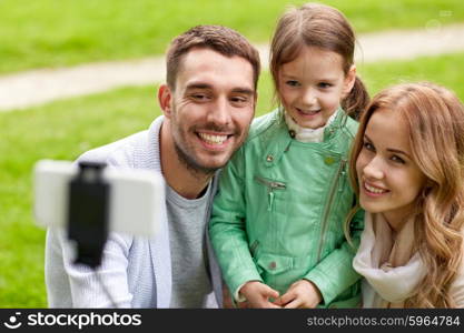 family, childhood, technology and people concept - happy father, mother and little daughter taking picture by smartphone selfie stick in park