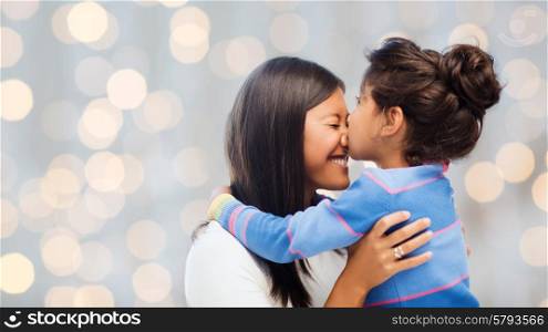 family, children and happy people concept - happy little girl hugging and kissing her mother over holidays lights background