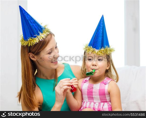 family, children, celebration and happy people concept - happy mother and daughter in blue party hats with favor horns