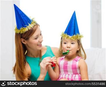 family, children, celebration and happy people concept - happy mother and daughter in blue party hats with favor horns