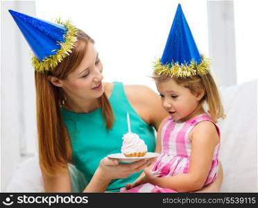family, children, celebration and happy people concept - happy mother and daughter in blue party hats with cake and candle
