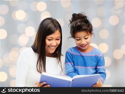family, children, education and happy people concept - happy mother and little daughter reading book over holidays lights background