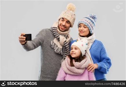 family, christmas and technology concept - happy mother, father and little daughter in winter clothes taking selfie by smartphone over grey background. happy family taking selfie by smartphone
