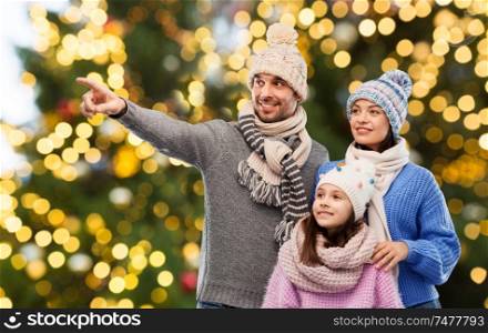 family, christmas and winter clothes concept - happy mother, father and little daughter in knitted hats and scarves over festive lights background. happy family in winter hats over christmas lights