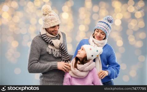family, christmas and winter clothes concept - happy mother, father and little daughter in knitted hats and scarves over festive lights background. family in winter clothes over christmas lights