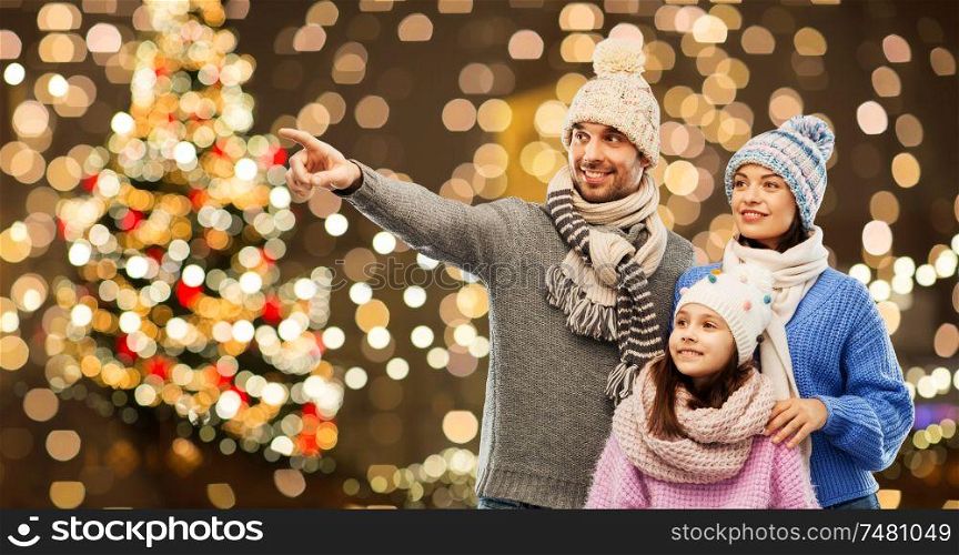 family, christmas and winter clothes concept - happy mother, father and little daughter in knitted hats and scarves over festive lights background. happy family in winter hats over christmas lights
