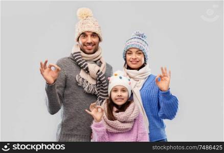 family, christmas and winter clothes concept - happy mother, father and little daughter in knitted hats and scarves showing ok gesture over grey background. happy family in winter clothes showing ok gesture