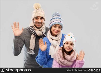 family, christmas and winter clothes concept - happy mother, father and little daughter in knitted hats and scarves waving hands over grey background. happy family in winter clothes waving hands