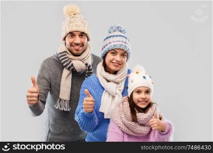 family, christmas and winter clothes concept - happy mother, father and little daughter in knitted hats and scarves showing thumbs up over grey background. happy family in winter clothes on grey background
