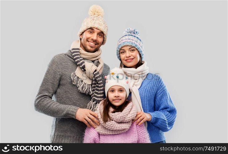 family, christmas and winter clothes concept - happy mother, father and little daughter in knitted hats and scarves over grey background. happy family in winter clothes on grey background