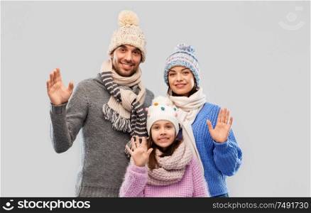family, christmas and winter clothes concept - happy mother, father and little daughter in knitted hats and scarves waving hands over grey background. happy family in winter clothes waving hands