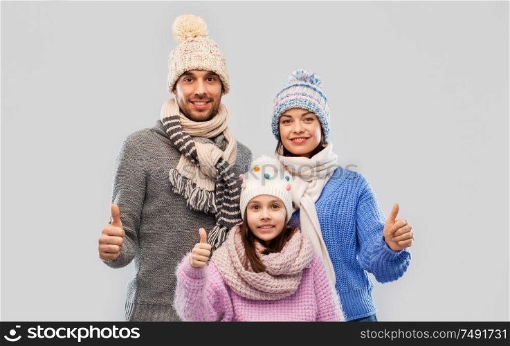 family, christmas and winter clothes concept - happy mother, father and little daughter in knitted hats and scarves showing thumbs up over grey background. happy family in winter clothes on grey background
