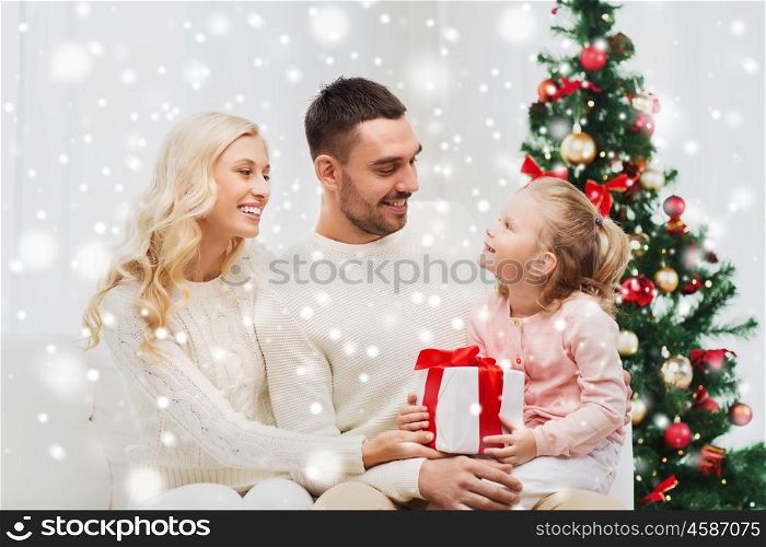 family, christmas, holidays and people concept - happy mother, father and little daughter with gift box sitting on sofa at home. happy family at home with christmas tree