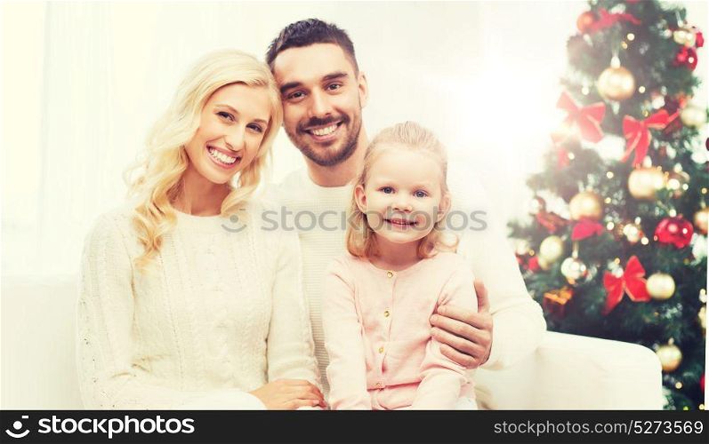 family, christmas, holidays and people concept - happy mother, father and little daughter sitting on sofa at home. happy family at home with christmas tree