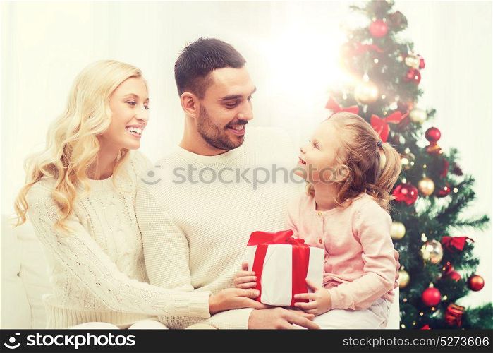 family, christmas, holidays and people concept - happy mother, father and little daughter with gift box sitting on sofa at home. happy family at home with christmas tree