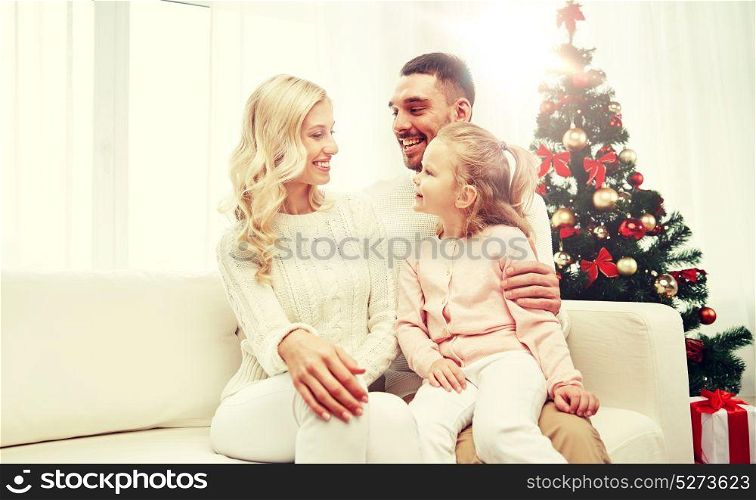 family, christmas, holidays and people concept - happy mother, father and little daughter sitting on sofa at home. happy family at home with christmas tree
