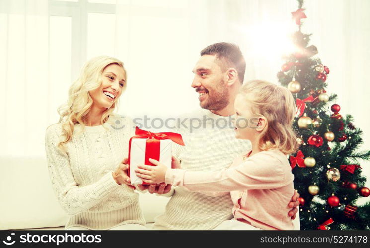 family, christmas, holidays and people concept - happy mother, father and little daughter with gift box sitting on sofa at home. happy family at home with christmas gift box