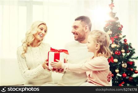 family, christmas, holidays and people concept - happy mother, father and little daughter with gift box sitting on sofa at home. happy family at home with christmas gift box