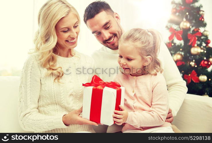 family, christmas, holidays and people concept - happy mother, father and little daughter with gift box sitting on sofa at home. happy family at home with christmas gift box