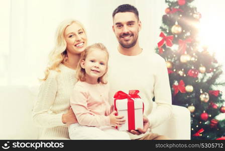 family, christmas, holidays and people concept - happy mother, father and little daughter with gift box sitting on sofa at home. happy family at home with christmas gift box