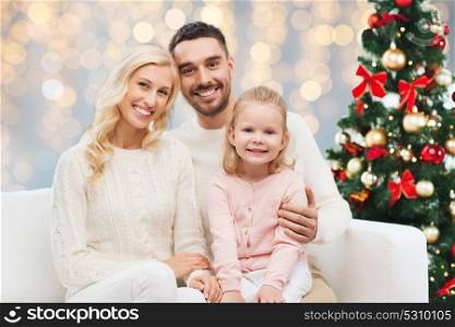 family, christmas, holidays and people concept - happy mother, father and little daughter sitting on sofa over lights background. happy family over christmas tree