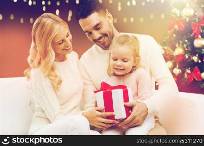 family, christmas, holidays and people concept - happy mother, father and little daughter with gift box sitting on sofa over lights background. happy family with christmas gift