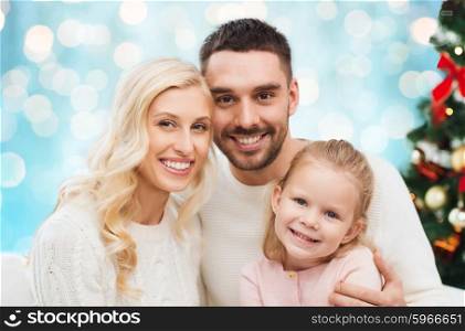family, christmas, holidays and people concept - happy mother, father and little daughter sitting on sofa at home over blue lights background