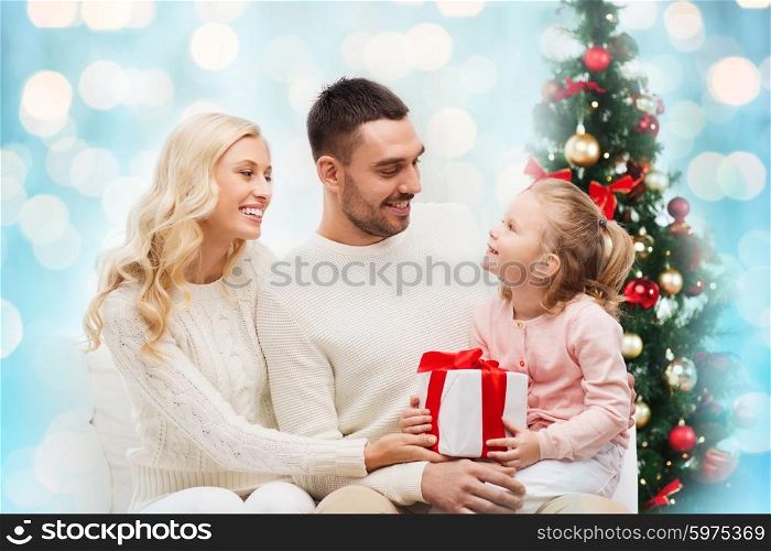 family, christmas, holidays and people concept - happy mother, father and little daughter with gift box sitting on sofa over blue holidays lights background