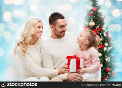 family, christmas, holidays and people concept - happy mother, father and little daughter with gift box sitting on sofa over blue holidays lights background