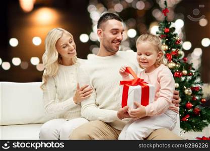 family, christmas, holidays and people concept - happy mother, father and little daughter with gift box sitting on sofa over lights background. happy family at home with christmas gift box