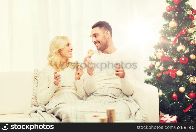 family, christmas, holidays, love and people concept - happy couple covered with plaid drinking tea and sitting on sofa at home. happy couple at home with christmas tree