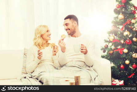 family, christmas, holidays, love and people concept - happy couple covered with plaid drinking tea and sitting on sofa at home. happy couple at home with christmas tree