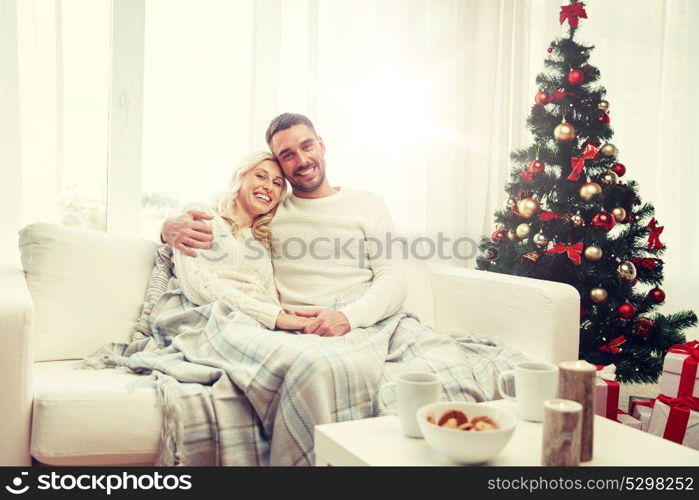 family, christmas, holidays, love and people concept - happy couple covered with plaid sitting on sofa at home. happy couple at home with christmas tree