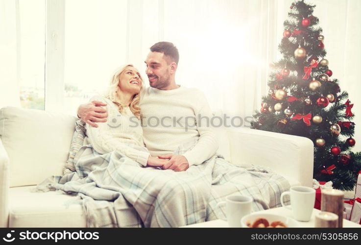 family, christmas, holidays, love and people concept - happy couple covered with plaid sitting on sofa at home. happy couple at home with christmas tree